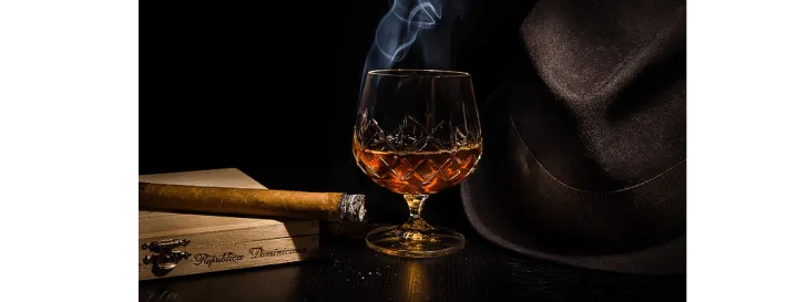 A smoking cigar on top of a wooden humidor, with a bit of ash on the end.  A glass of brandy, and a brown fedora.